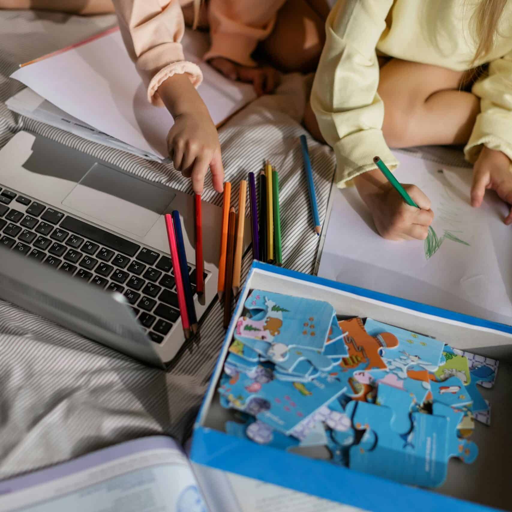 Two children drawing and solving puzzles at home, surrounded by colored pencils and a laptop.