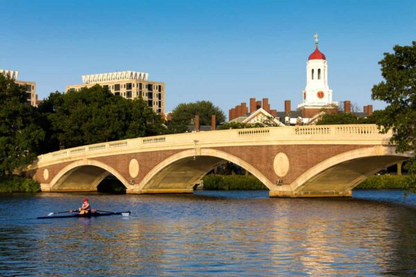 Rower on the Charles River passing under the historic Anderson Memorial Bridge in Cambridge, Massachusetts, with Harvard University buildings in the background.