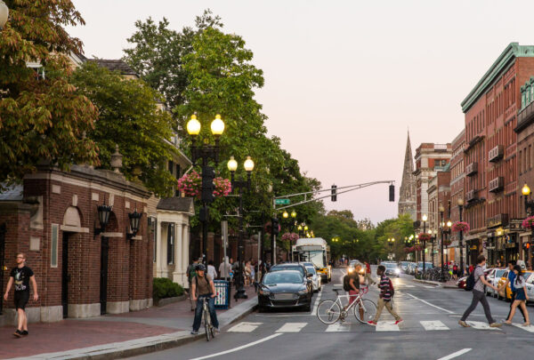Pedestrians and cyclists crossing a busy street in Harvard Square, Cambridge, Massachusetts, during the evening with cars, shops, and historic buildings in the background.