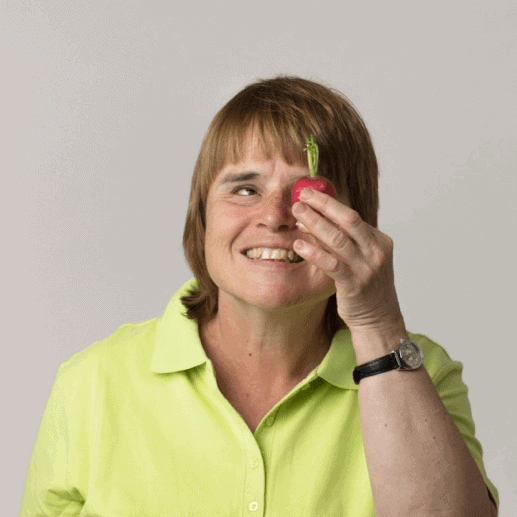 Ein Profilbild von Nina Schweppe. Sie hält sich mit ihrer linken Hand ein Radieschen vor ihr linkes Auge und lächelt in die Kamera.