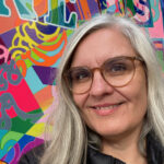 Yvonne Frank lacht in die Kamera. Sie befindet sich auf der Ausstellung NordArt und steht vor einem farbenfrohen Kunstwerk mit dem Namen „Happiness is a Choice“. Yvonne hat graue Haare und trägt eine schwarze Jacke.