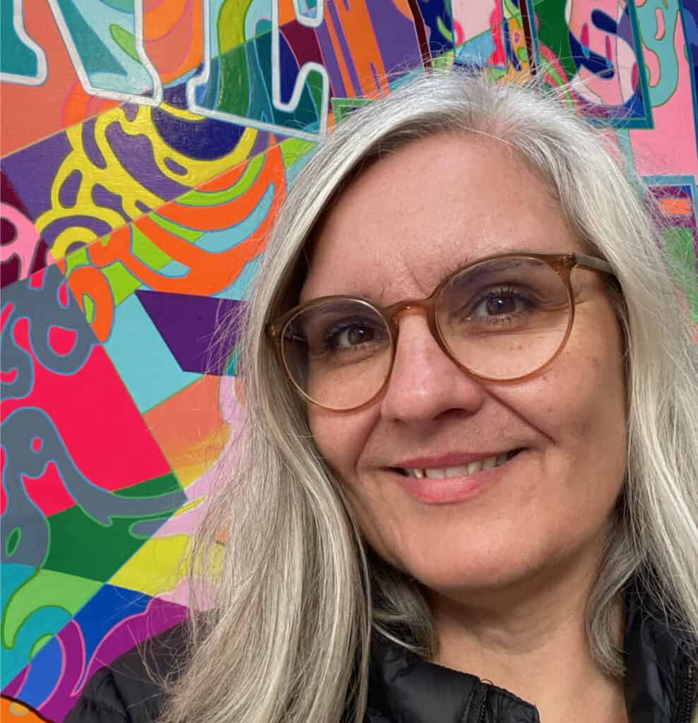Yvonne Frank lacht in die Kamera. Sie befindet sich auf der Ausstellung NordArt und steht vor einem farbenfrohen Kunstwerk mit dem Namen „Happiness is a Choice“. Yvonne hat graue Haare und trägt eine schwarze Jacke.