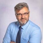 Dirk Haas trägt ein blaues Hemd und eine dunkelblaue Krawatte. Er hat eine dünne, schwarz-silberne Brille und graue, kurze Haare.