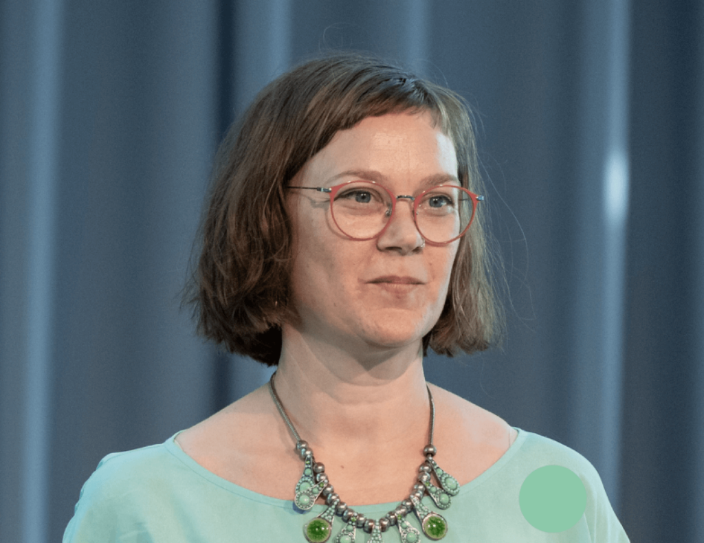 Inga Schiffler lächelt eine Kamera. Sie hat kinnlange, braune Haare, eine Brille, ein grünes Oberteil und eine farblich passende Kette mit grünen Steinen.