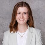 Profilfoto von Marie Stroh. Sie trägt schulterlange Haare, einen grauen Blazer über ein weißes Shirt und lächelt dich an