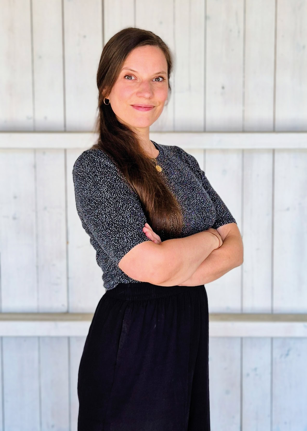 Christiane steht mit verschränkten Armen freundlich lächelnd nach rechts geneigt vor einer weißen Holzwand. Sie hat langes braunes Haar und trägt ein schwarz weiß gepunktetes Shirt.