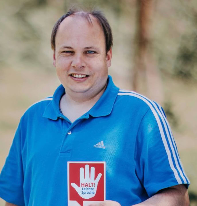 Mario Stabel hat ein blaues Polo-Shirt an. In der Hand hält ein Schild auf dem steht: Halt! Leichte Sprache.