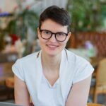 Olivia lächelt in die Kamera. Sie hat schwarze kurze Haare, eine schwarze Brille und trägt ein weißes T-Shirt.