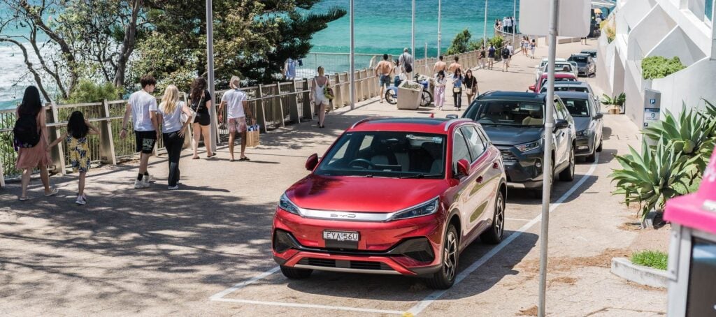 Modern red SUV parked near beach with pedestrians and ocean view in background.
