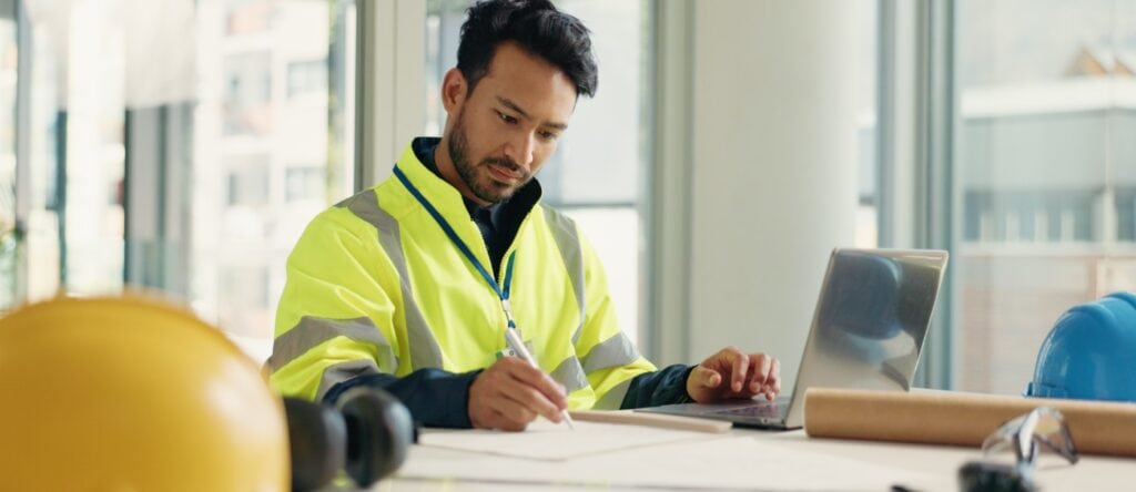 High-visibility worker in safety gear reviewing plans at construction site office.