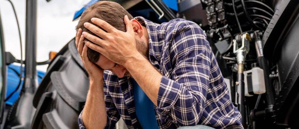 Struggling male mechanic holding his head in front of engine, feeling stressed or overwhelmed.