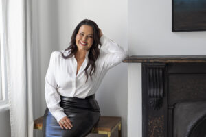 A woman smiling in a white blouse and black skirt sitting near a fireplace.