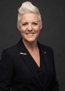 Elegant senior woman with short, styled blonde hair wearing a black blazer against a dark background.
