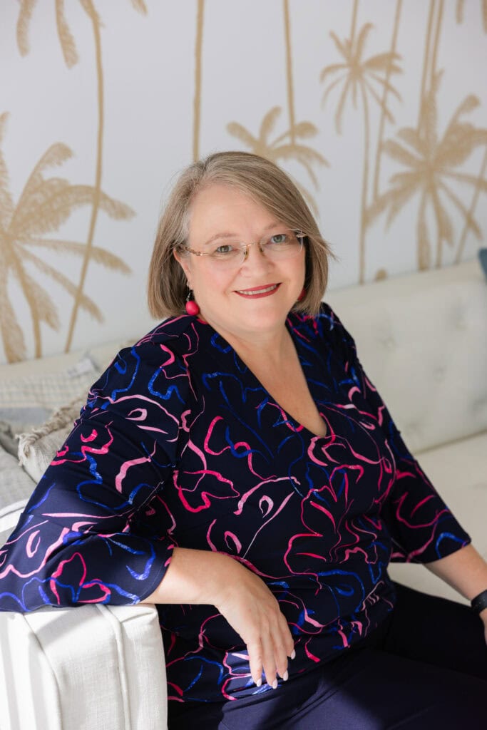 A woman smiling and sitting on a white sofa in a modern, stylish room with palm tree wall art.