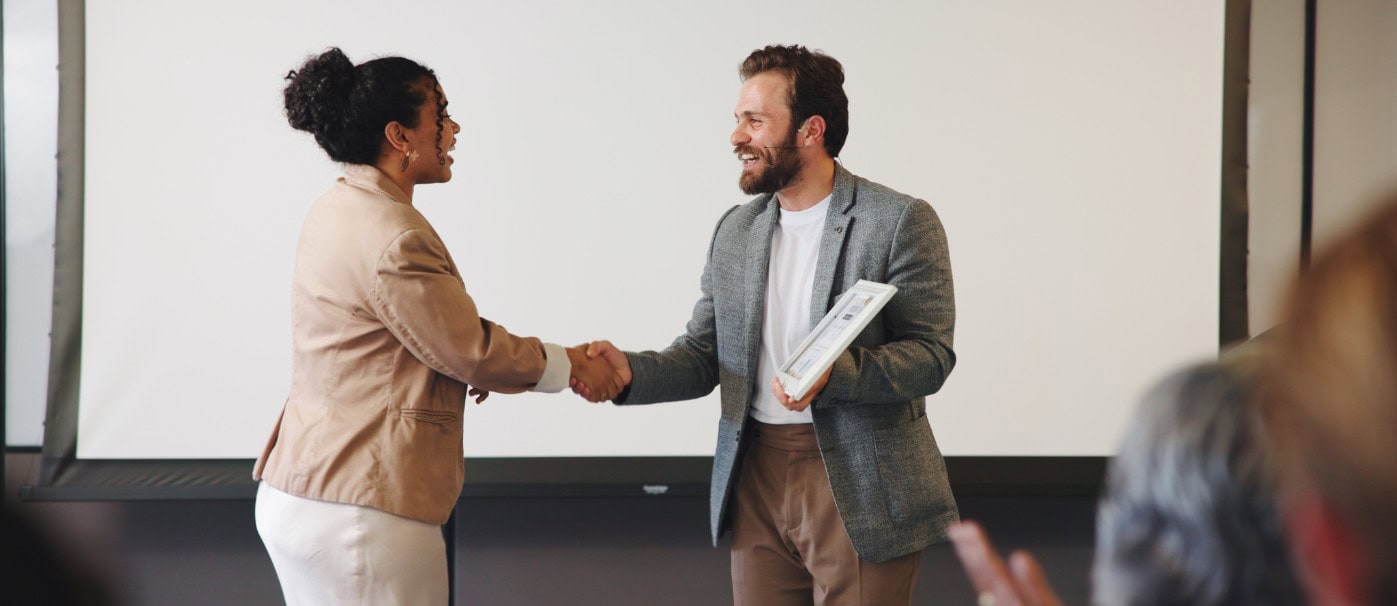 A professional handshake between a woman and a man in a business setting, with a presentation screen.