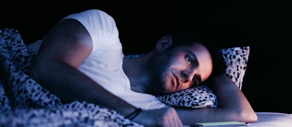 Man lying in bed using a tablet at night, dark bedroom setting.