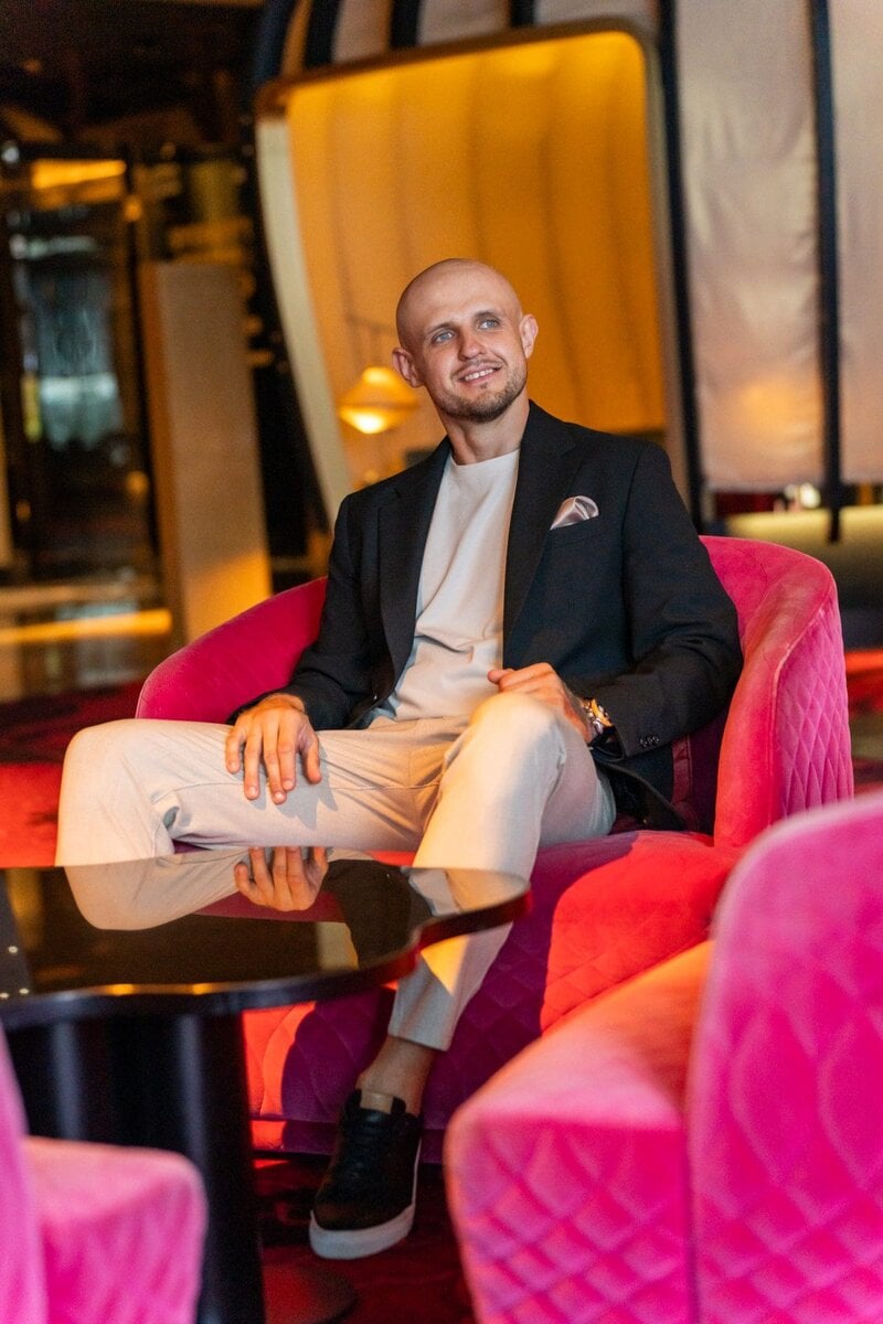 Int: Young bald man in a dark jacket sitting in a pink seat having his dating photo taken by dating photographer Georgie Green in Sydney NSW