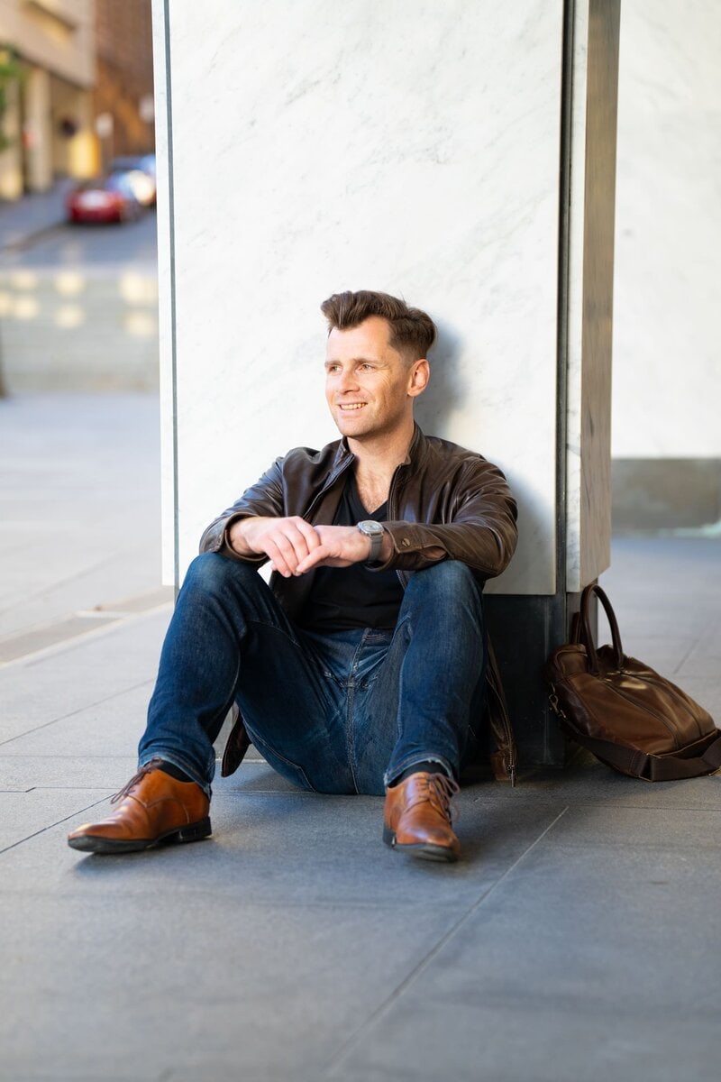 Young dark haired man wearing a brown leather jacket sitting by a street pillar having his dating photo taken by Sydney dating photographer Georgie Greene