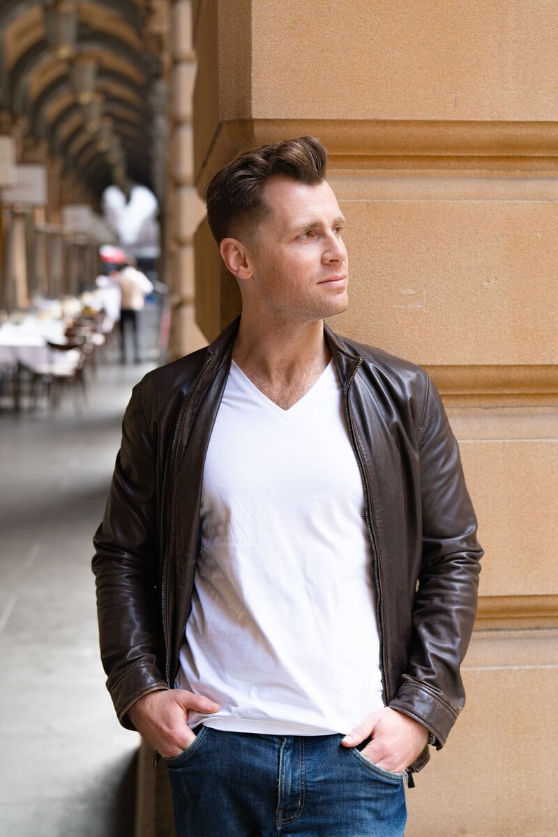 Young dark haired man wearing a brown leather jacket standing by a street pillar having his dating photo taken by Sydney dating photographer Georgie Greene