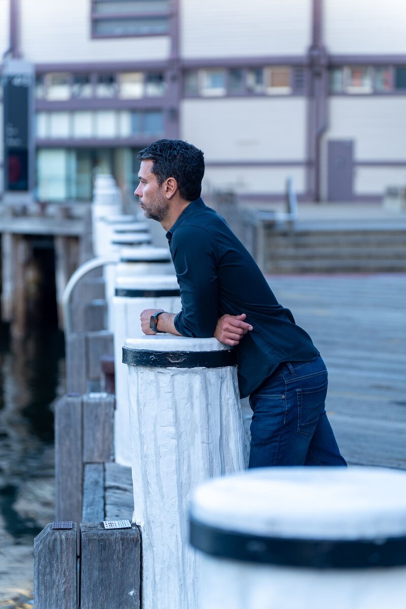 Dark hair man with beard wearing a blue shirt and leaning on a post having his dating photo taken by top Sydney dating photographer Georgie