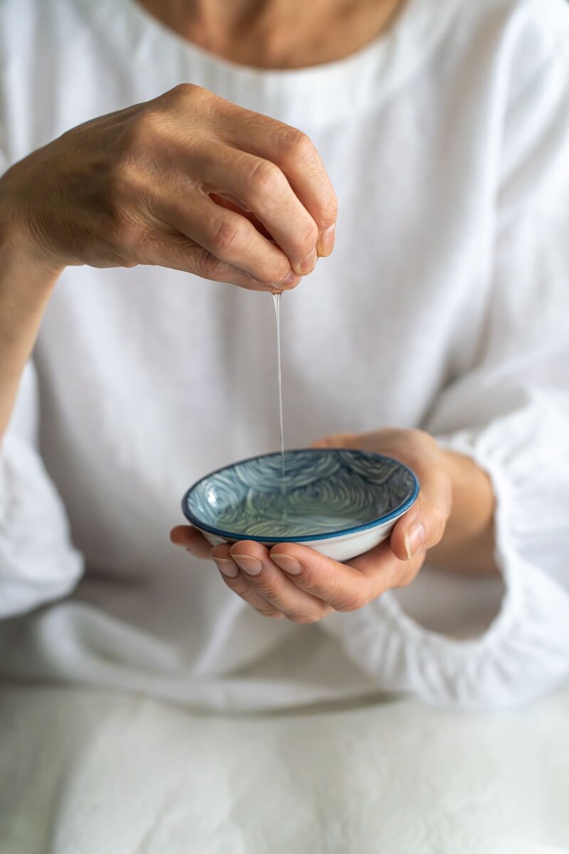 Hand holding a drop of oil as it flows into a small dish below