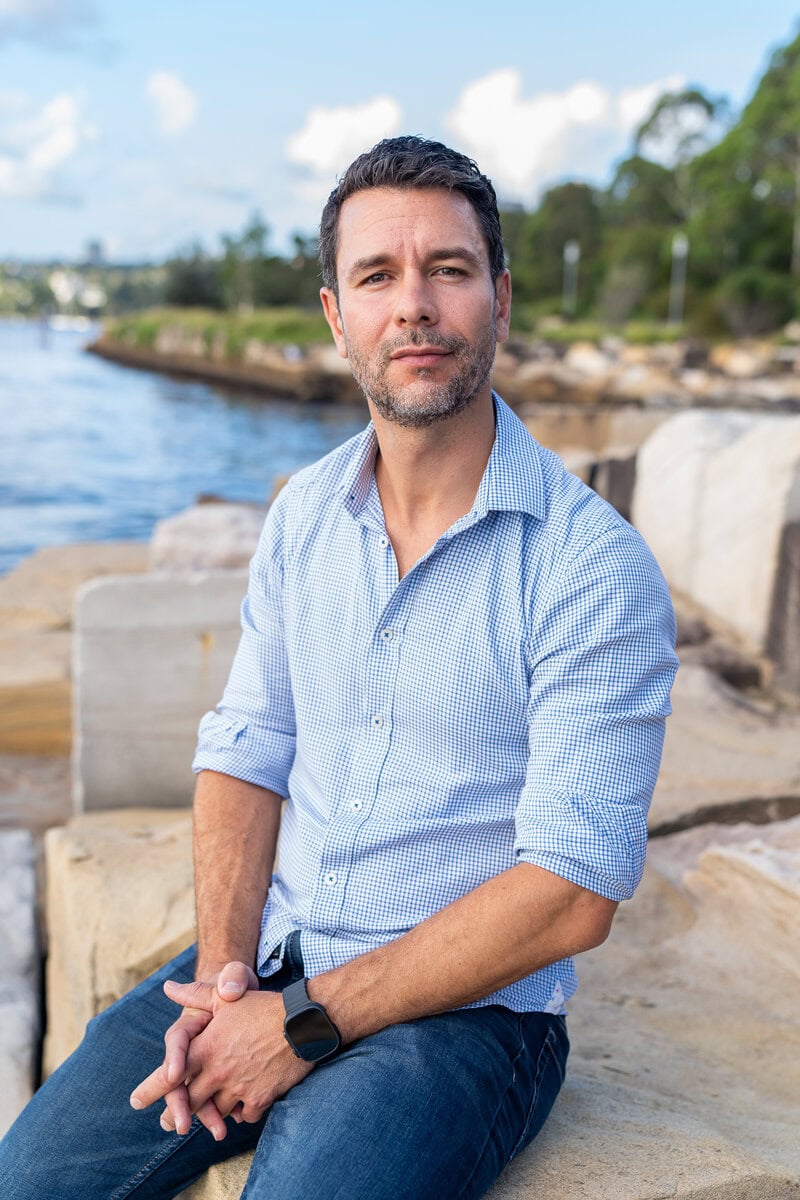 Dark hair man with beard wearing a blue shirt and sitting by the water having his dating photo taken by top Sydney dating