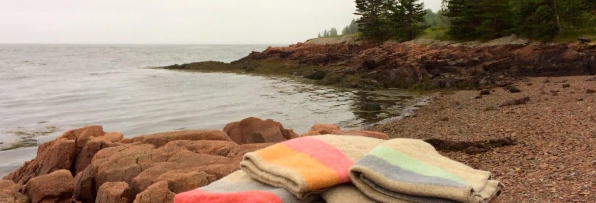 Soft pastel-colored wool blankets, featuring rainbow stripes, laid on rocks by a lakeside with trees and cliffs, highlighting Seacolors Yarnery's natural and vibrant yarns collection.