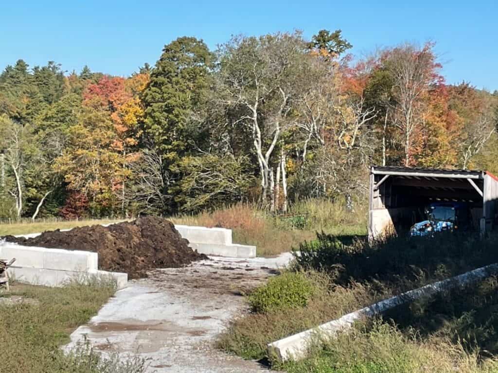 Colorful autumn trees with fall foliage in background, construction site with concrete blocks and dirt pile, and a shed with a vehicle inside, showcasing rural outdoor scene for SeaColors Yarnery farm-inspired craft setting.