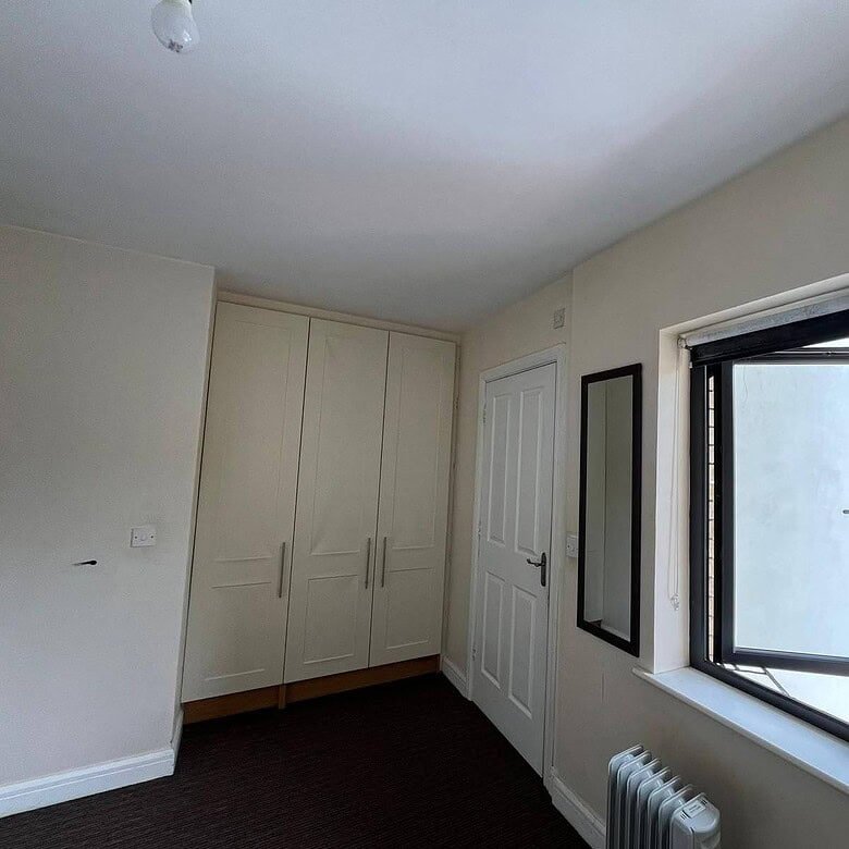 Double room with ensuite in Dublin 22 featuring a wardrobe, window, radiator, and door. – Ghar.ie property listing