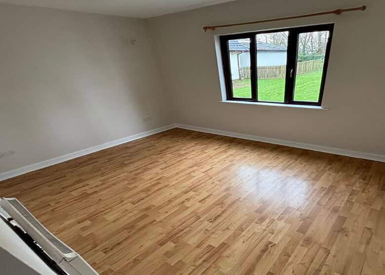 Empty bedroom with wood laminate flooring, cream walls, and a window view of a green lawn and white building.