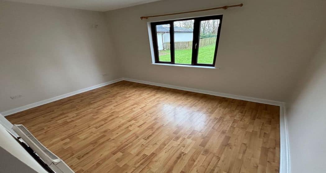 Empty bedroom with wood laminate flooring, cream walls, and a window view of a green lawn and white building.