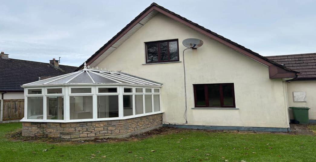House with conservatory addition, stone base, white frame, dark windows, satellite dish, and green lawn.