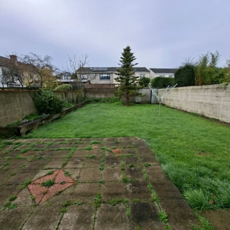 Backyard with grass, patio, tree, and concrete walls under cloudy sky.