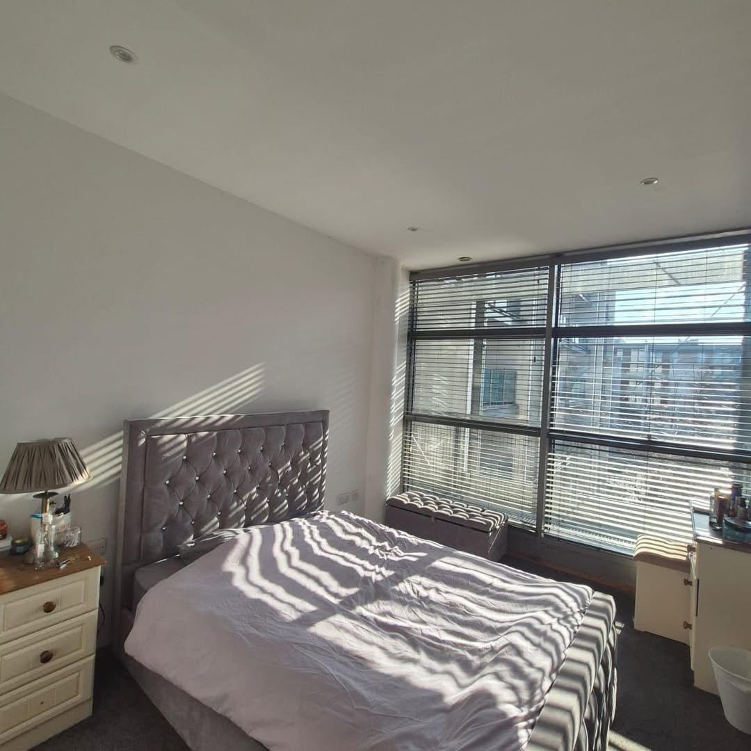 Bedroom with grey bed, tufted headboard, and sunlight streaming through blinds.