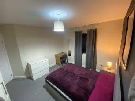 Bedroom with purple bedding, desk, and white dresser. Neutral walls and carpet. Modern lighting.