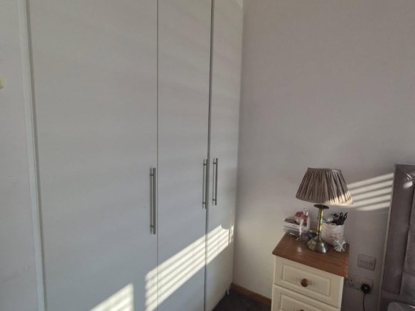 Bedroom corner with white wardrobe, nightstand, and lamp. Sunlight through blinds.