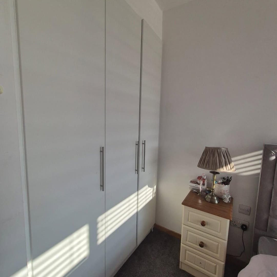 Bedroom corner with white wardrobe, nightstand, and lamp. Sunlight through blinds.