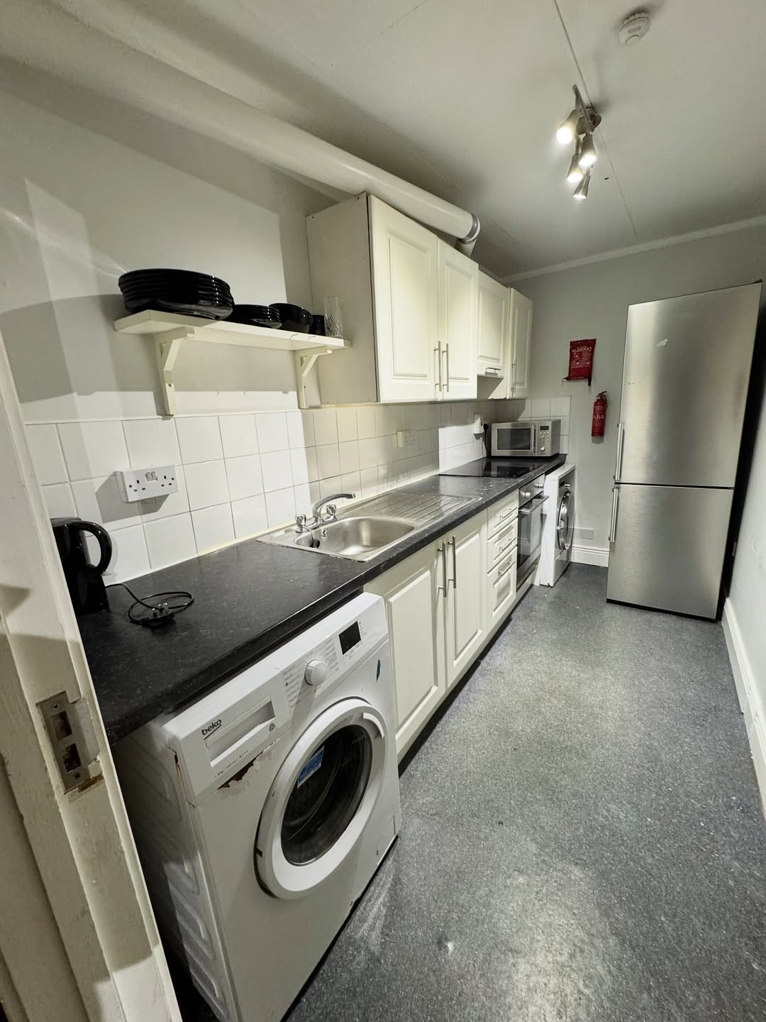 Compact kitchen with white cabinets, black countertop, stainless steel fridge, and washing machine.