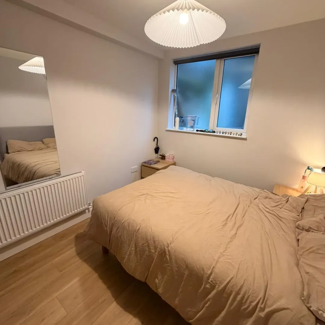Cozy bedroom with double bed, radiator, and window. Neutral tones, warm lighting.