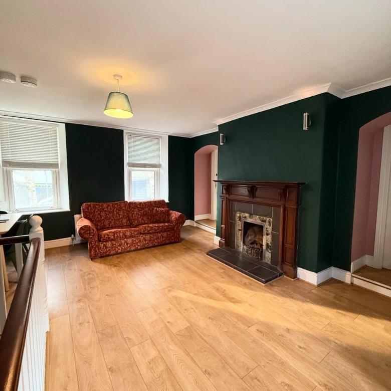 Living room with dark green walls, fireplace, red sofa, and light wood floors. Windows with blinds.