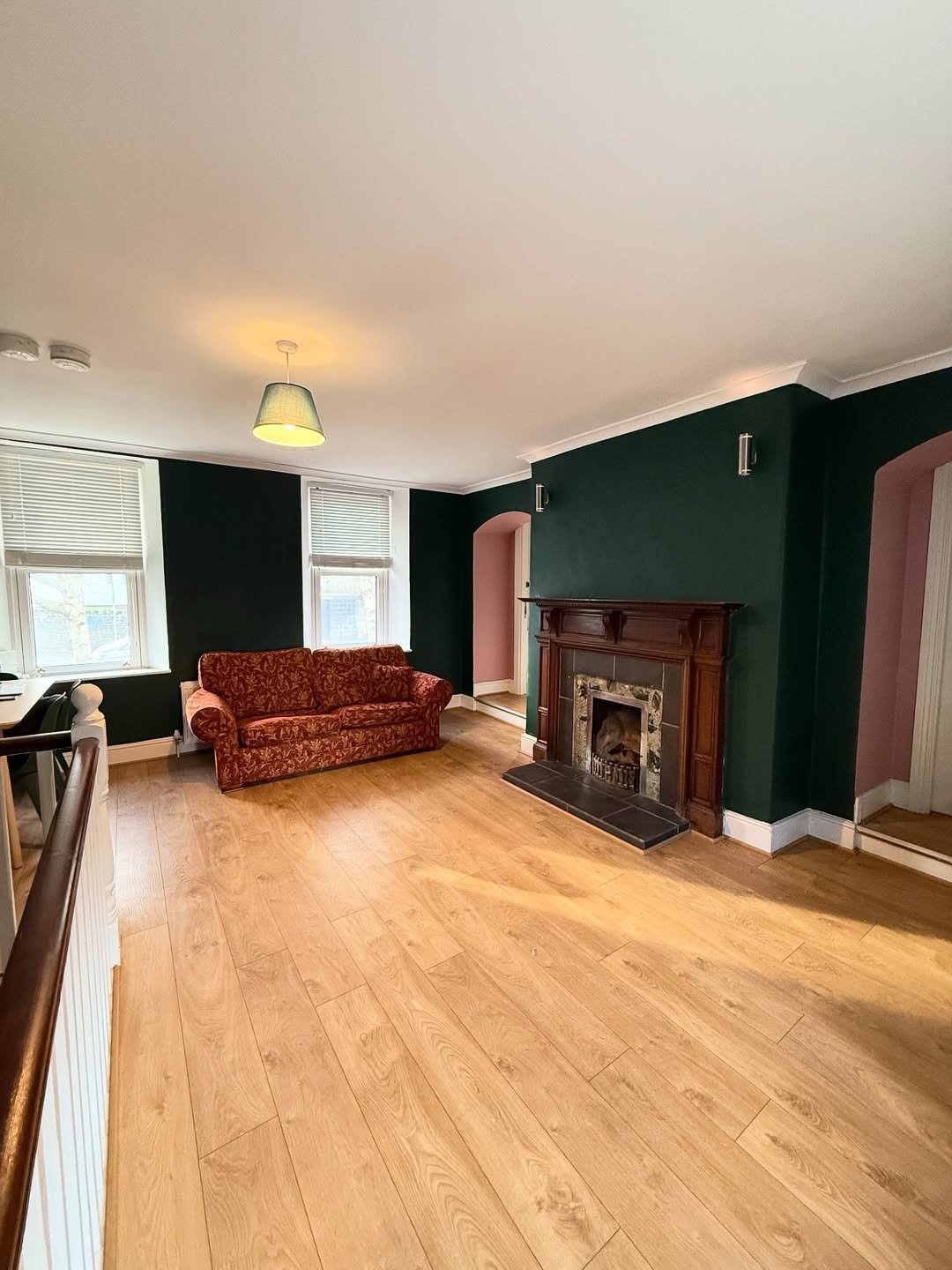 Living room with dark green walls, fireplace, red sofa, and light wood floors. Windows with blinds.