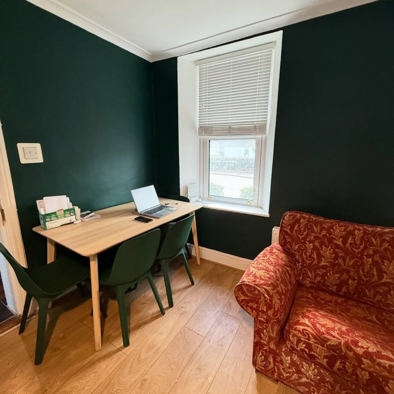 Home office setup with desk, laptop, green chairs, and red armchair near window.