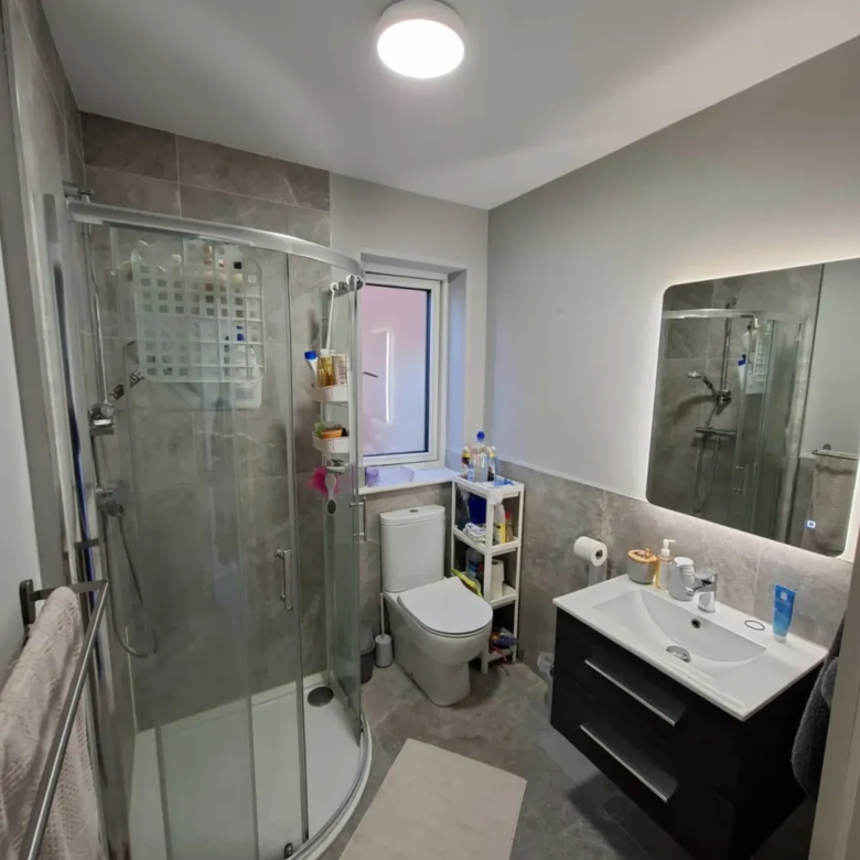 Modern bathroom with shower, toilet, vanity, and illuminated mirror. Gray tile and white walls.