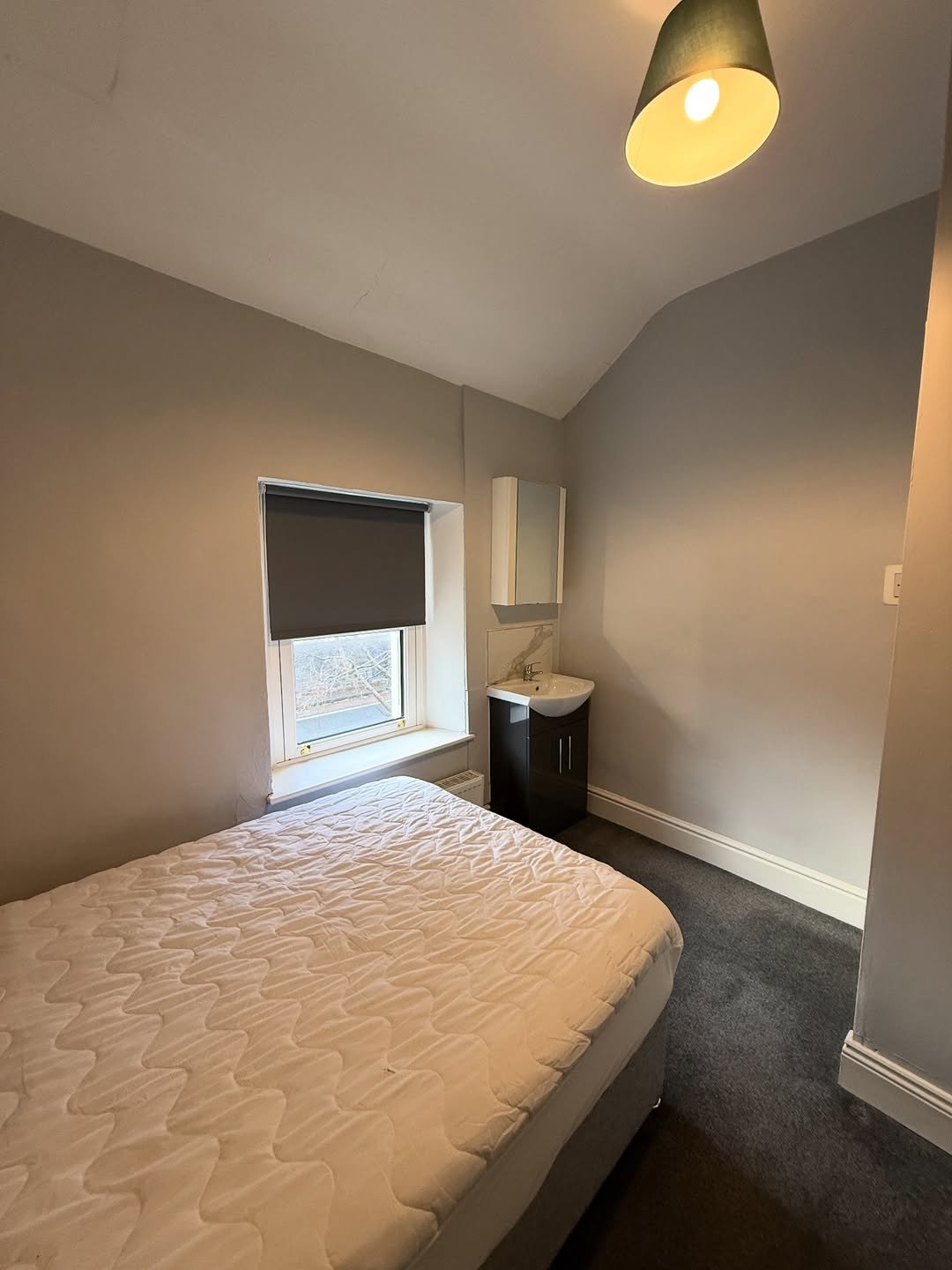 Small bedroom with bed, sink, cabinet, window with blind, gray walls, and dark carpet.