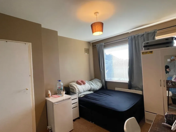 Cozy bedroom with bed, wardrobe, desk, and small refrigerator. Natural light from window.