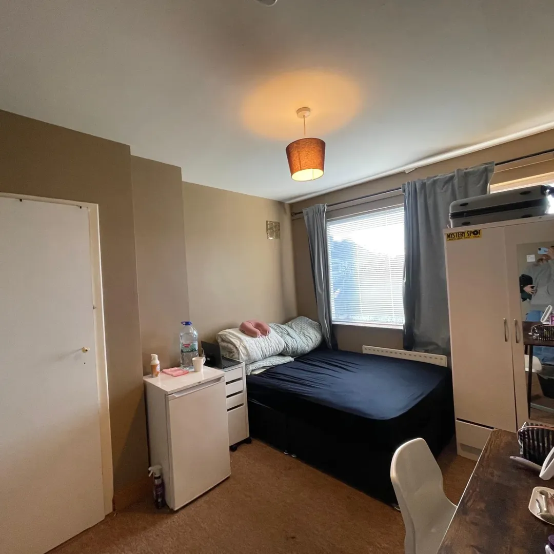 Cozy bedroom with bed, wardrobe, desk, and small refrigerator. Natural light from window.