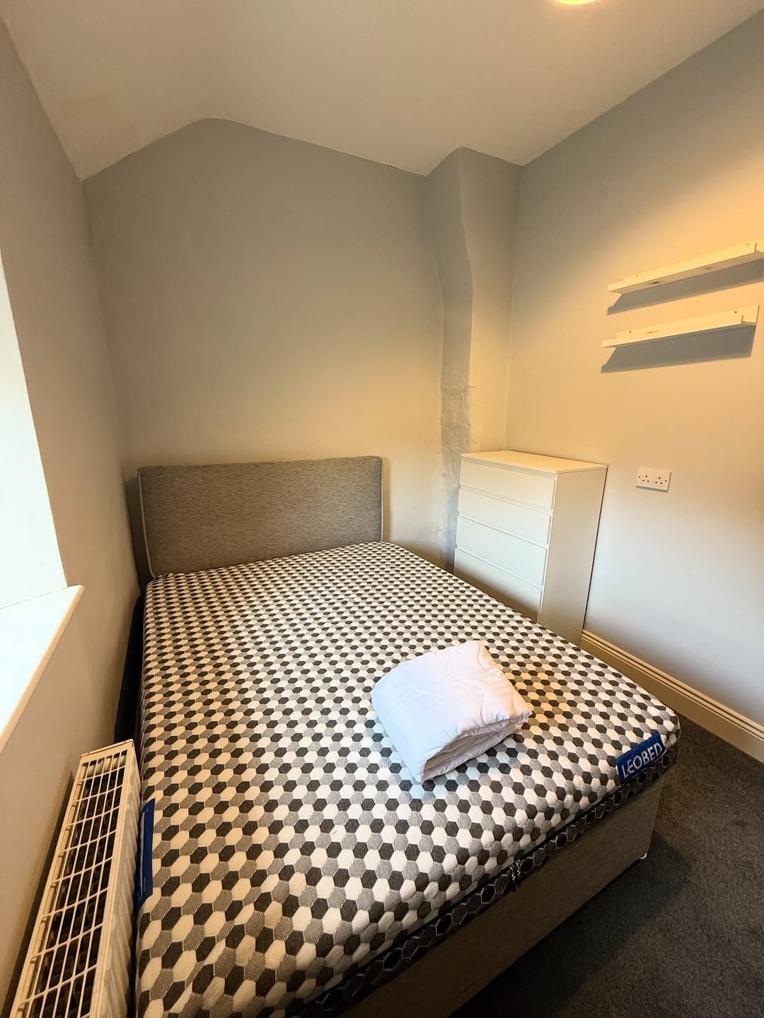 Cozy bedroom with patterned bed, white dresser, and radiator. Simple room interior.