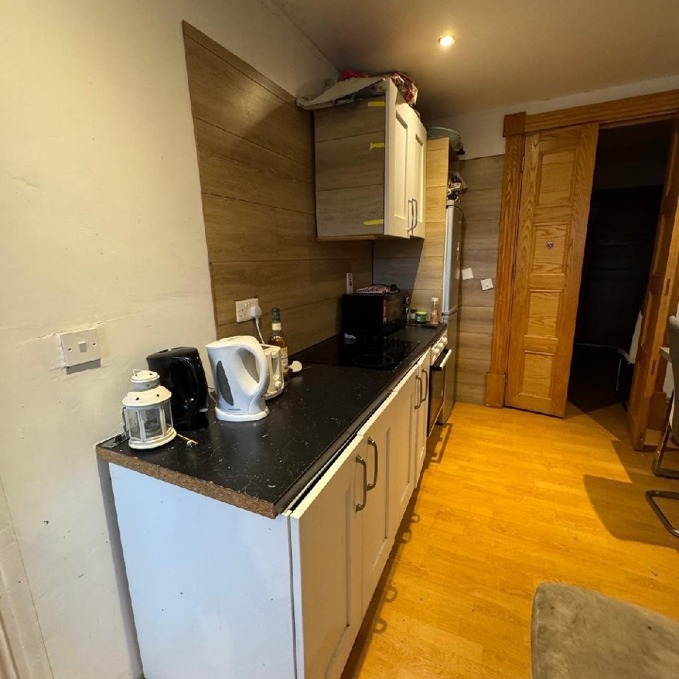 Compact kitchen with white cabinets, black countertop, kettle, and doorway.