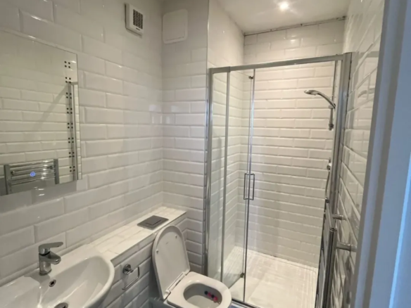Small white tiled bathroom with shower, toilet, and sink.