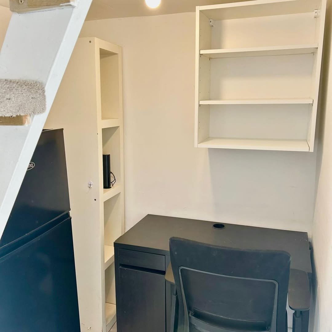 Compact workspace: black desk, chair, shelves, and mini fridge under stairs.